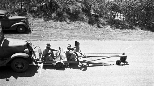 Two men seated on machine used for painting center line stripe on road in Zion Canyon. Machine is mounted to front of truck. Vehicle parked on shoulder in background. [Arrows drawn onto image]