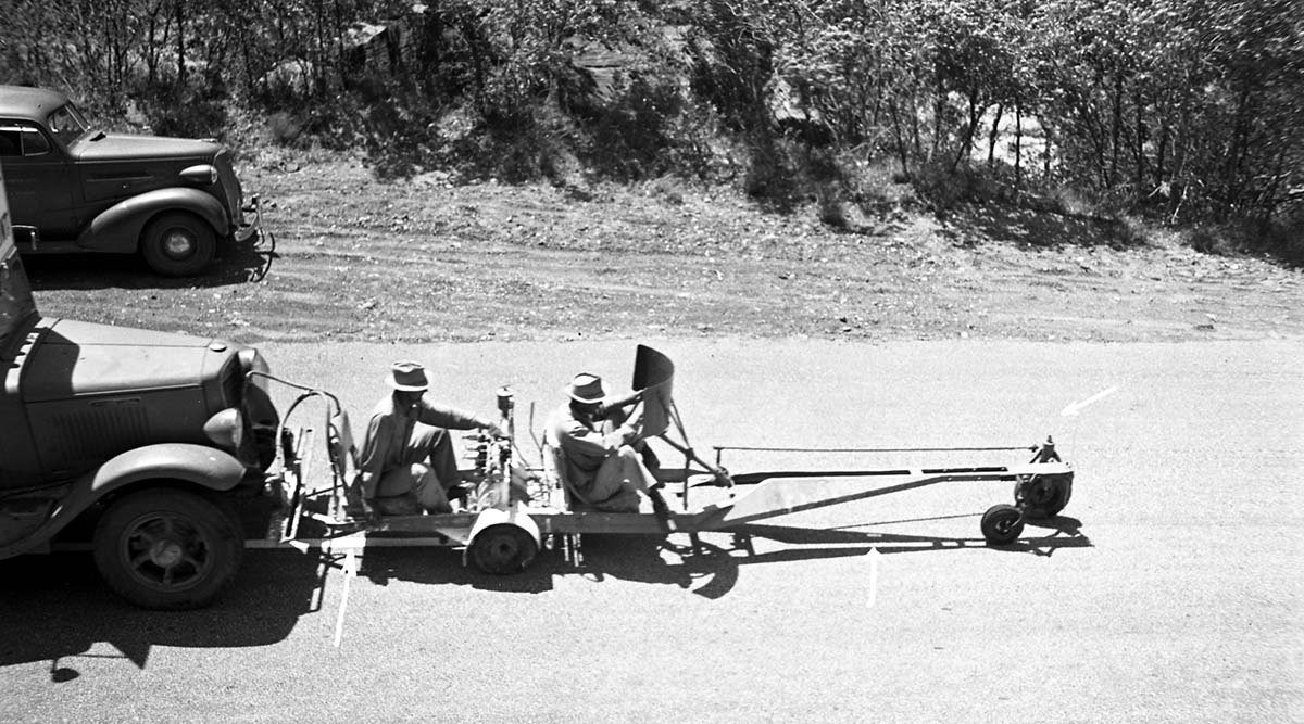 Two men seated on machine used for painting center line stripe on road in Zion Canyon. Machine is mounted to front of truck. Vehicle parked on shoulder in background. [Arrows drawn onto image]