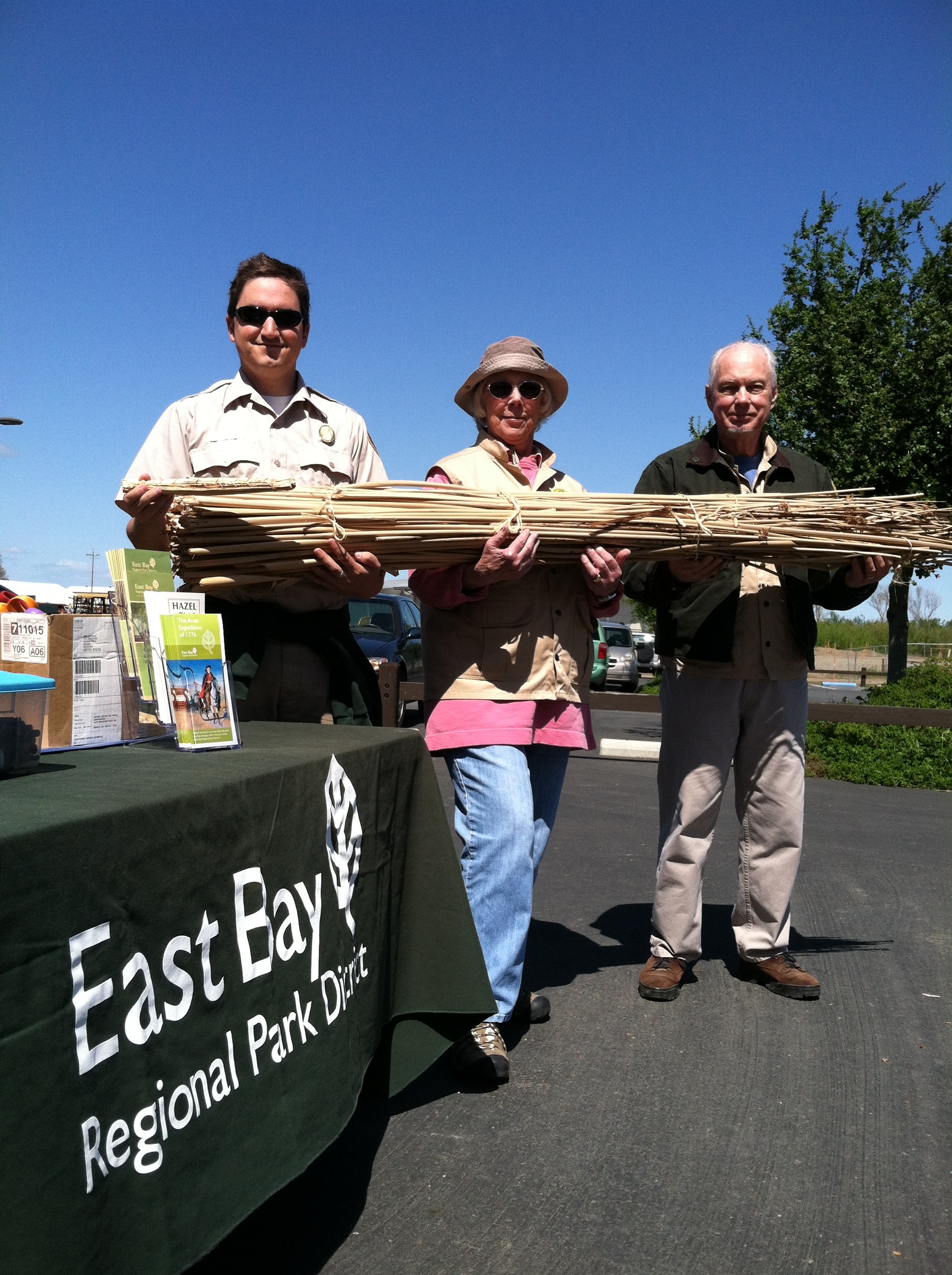 Three people stand in a row holding a large bundle of sticks next to a table with brochures