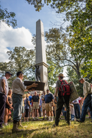 A park ranger wearing a red backpack talks to a crowd near a monument