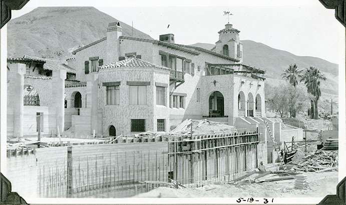 This is an historic black and white photograph from the Scotty's Castle Historic Photograph Collection, Death Valley National Park of Scotty's Castle Swimming Pool, west end looking northeast. Walls formed. Two laborers bottom right and Main House in background. May 18, 1931. Photographed by Mat Roy Thompson.