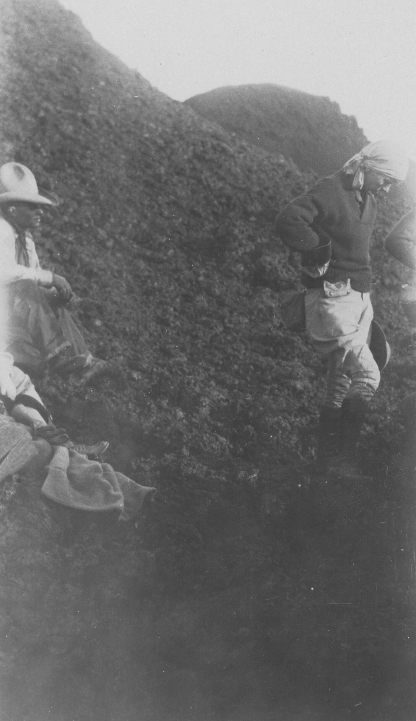 Black and white photograph of four people sitting and standing on a rocky slope. Only two people are fully visible. On the left side, a foot and hand are seen, assumed to be a person sitting down off to the left. Above this, another person is mostly visible, sitting with their arm resting on their knee, looking towards the right side of the image. A third person is fully visible on the right side. They are mid-step, looking down at the ground. A fourth person’s elbow is seen at the right edge of the photo.