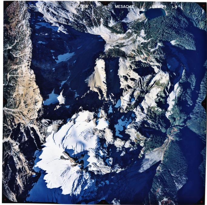 Color aerial photograph of glaciated mountain peak with forested valleys below.