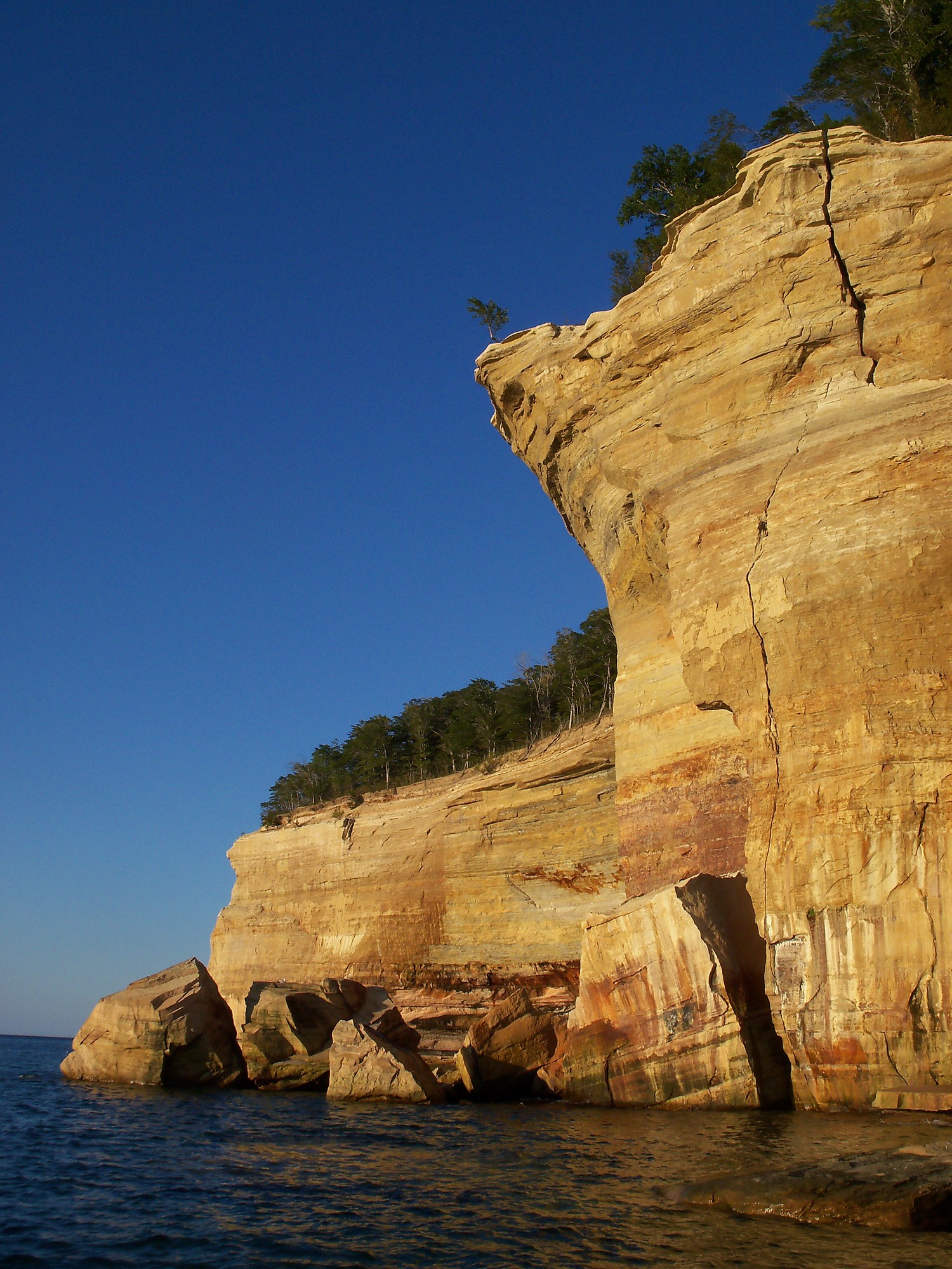 Pictured Rocks Cliffs are streaked with reds and oranges and black colors. These are caused by minerals seeping through the sandstone rock. A crack is visible stretching from the top to the bottom of a cliff section. Broken pieces of the cliff are at the base of the vertical wall of rock.