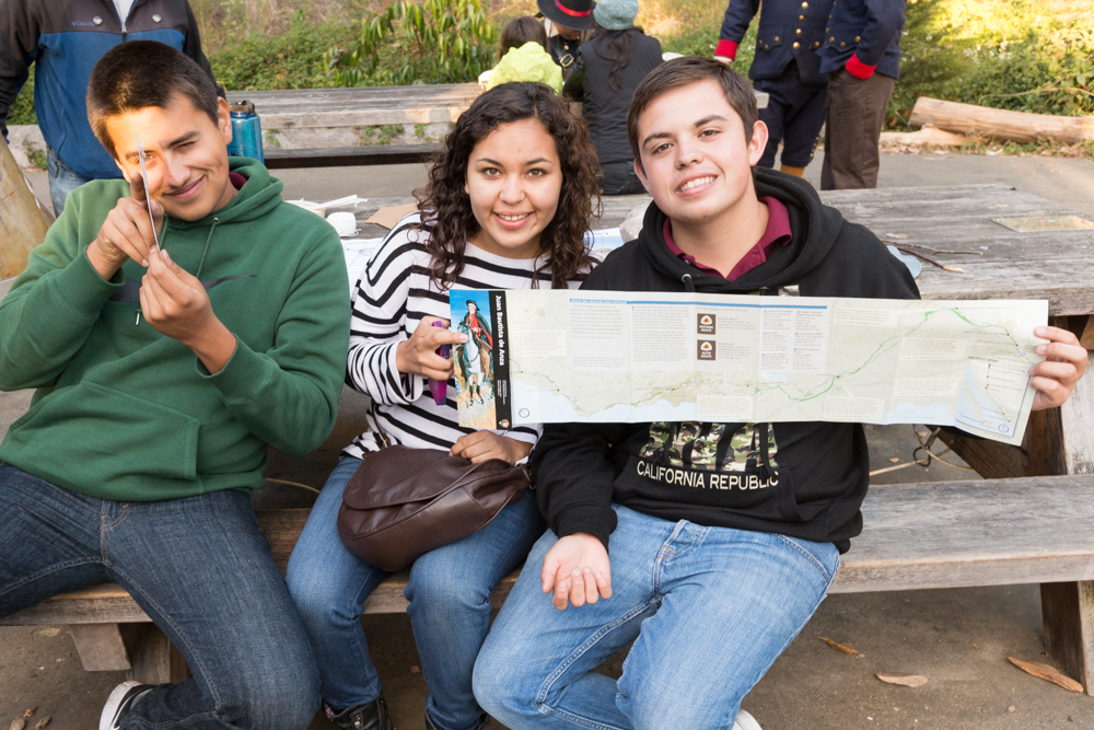 Three teenagers sit on a wooden picnic table bench holding the Juan Bautista de Anza brochure map