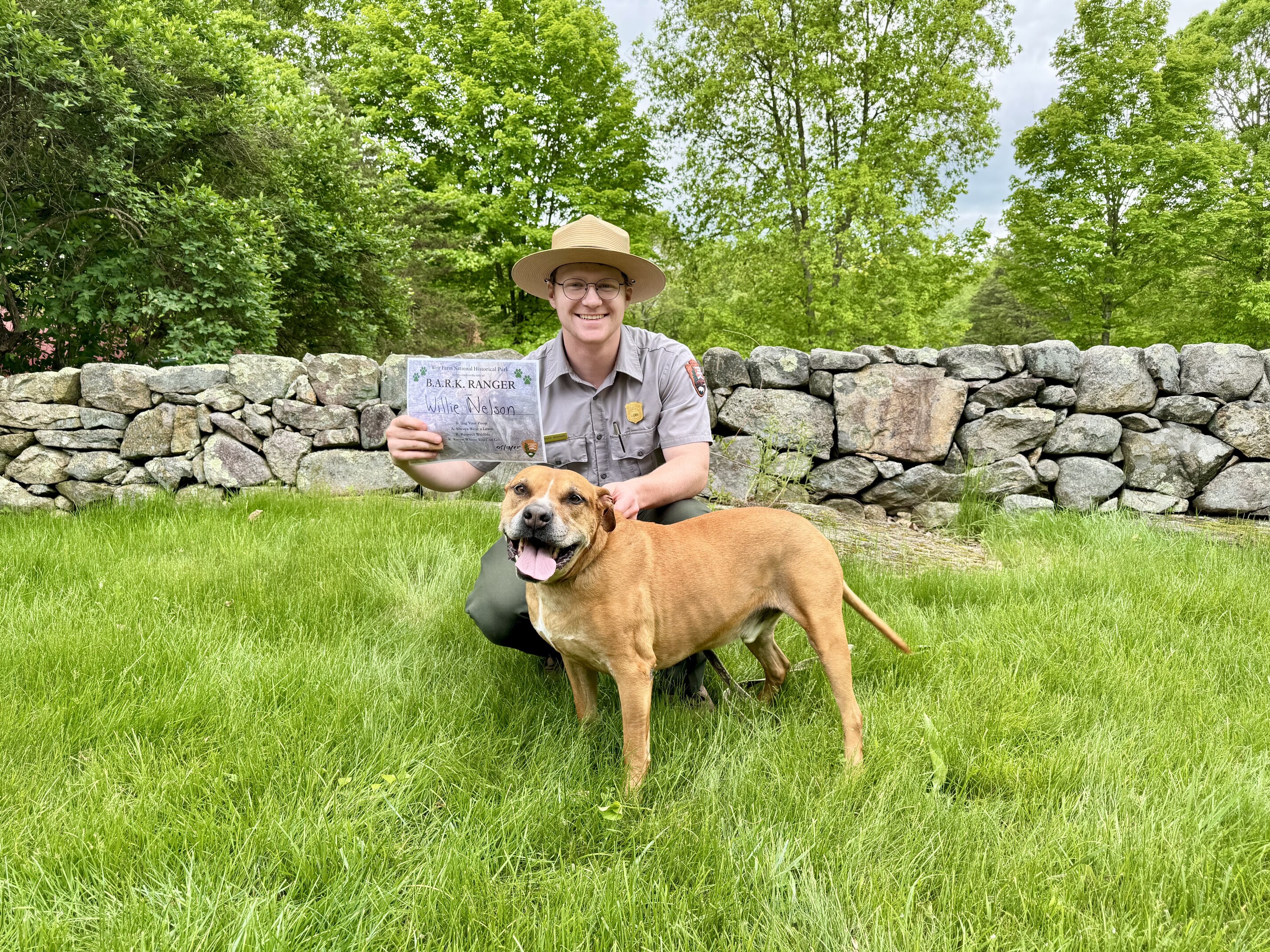 A person kneels in front of a stone wall holding a certificate behind a standing dog.