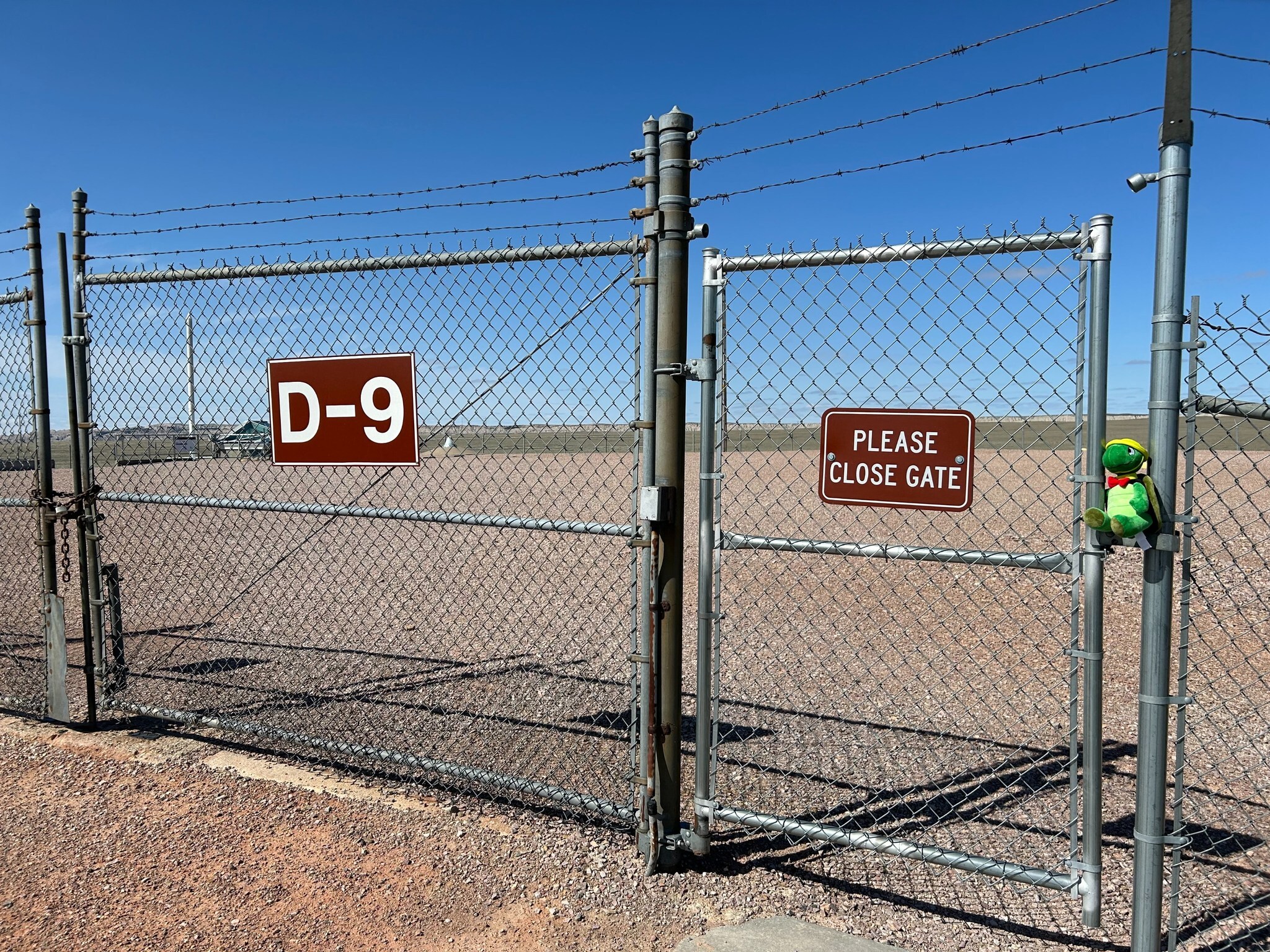 About an acre of gravel surrounded by a chain link fence. Signs for “D-9” and a reminder to “please close gate” hang from the chain link. 