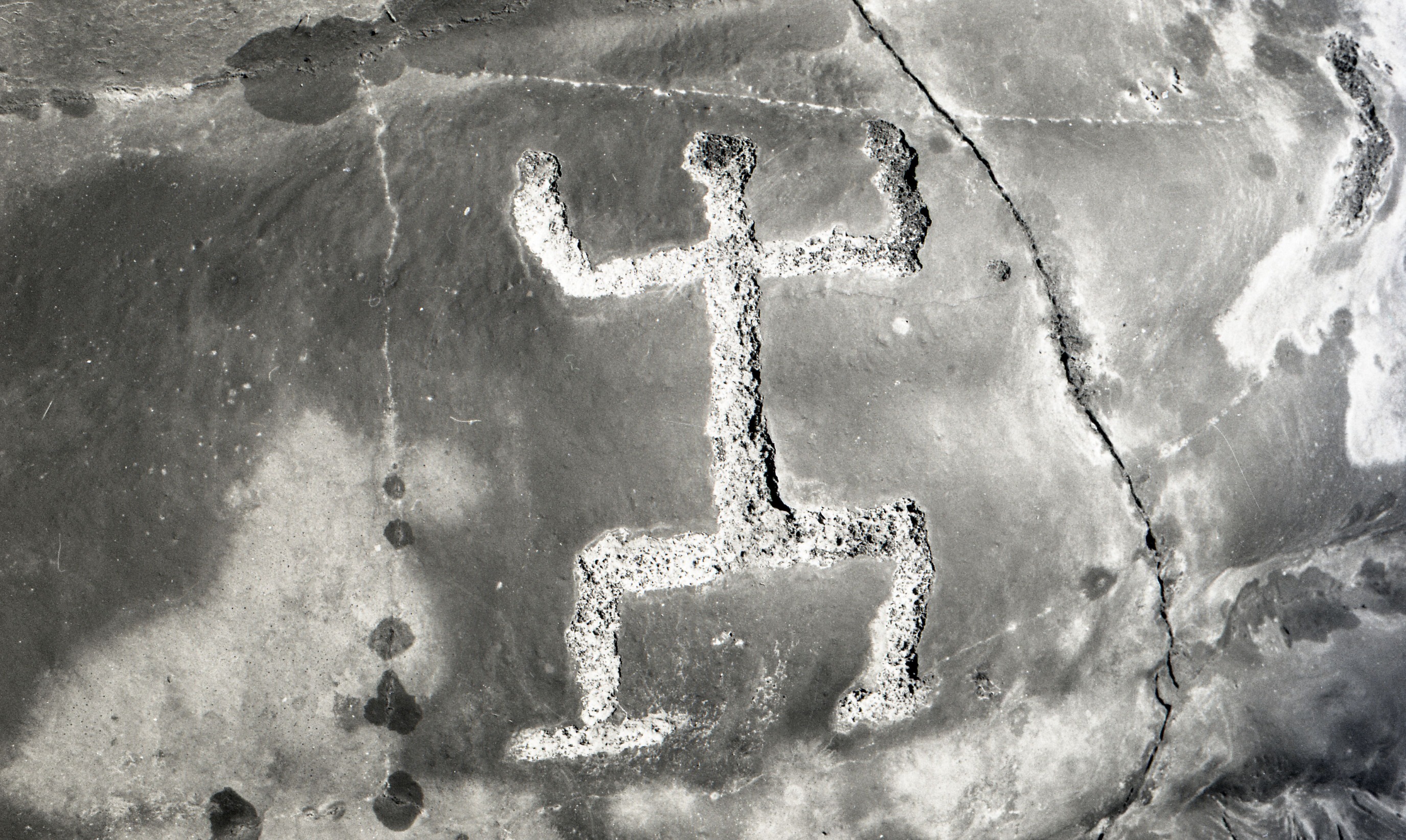A black and white image of a human-figured petroglyph carved onto a large smooth rock.
