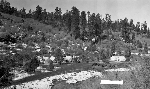 The Civilian Conservation Corps (CCC) spike camp (temporary camp sometimes referred to as a stub camp) located in Jolley Wash, viewed from a small rise. Visible in the photograph are seven tents in the wash among the pinyon and juniper trees, one small wood frame structure and a pickup truck. Ponderosa pine trees on the ridge in back, and a small amount of snow is visible on the ground. Civilian Conservation Corps (CCC) workers constructing the East Rim Trail were temporarily housed at the Jolley Wash campsite.