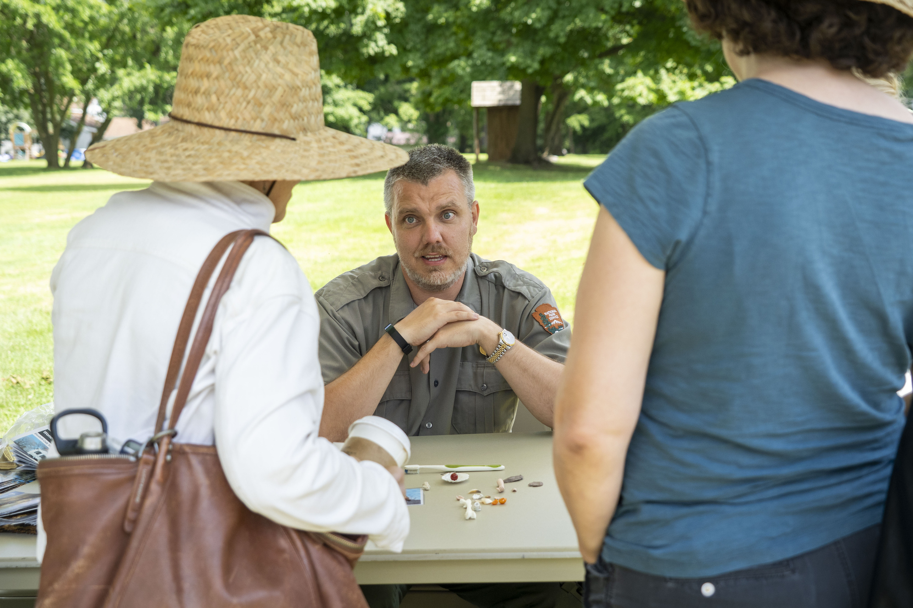 A man sits behind a table outdoors and speaks to two women across sthe table