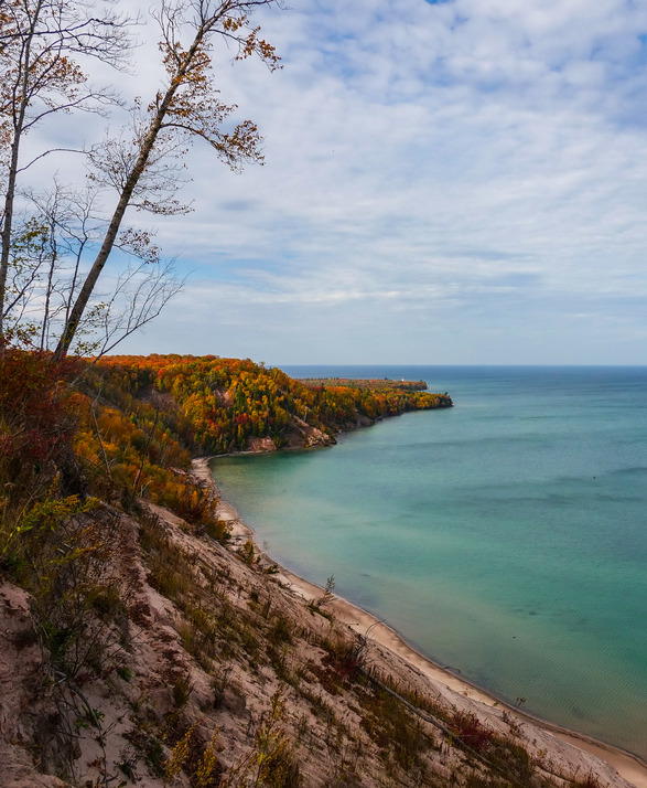 A peninsula juts out into Lake Superior. It is covered in trees in shades of green, yellow, and red. A white and black lighthouse tower pokes out of the trees in the distance. 