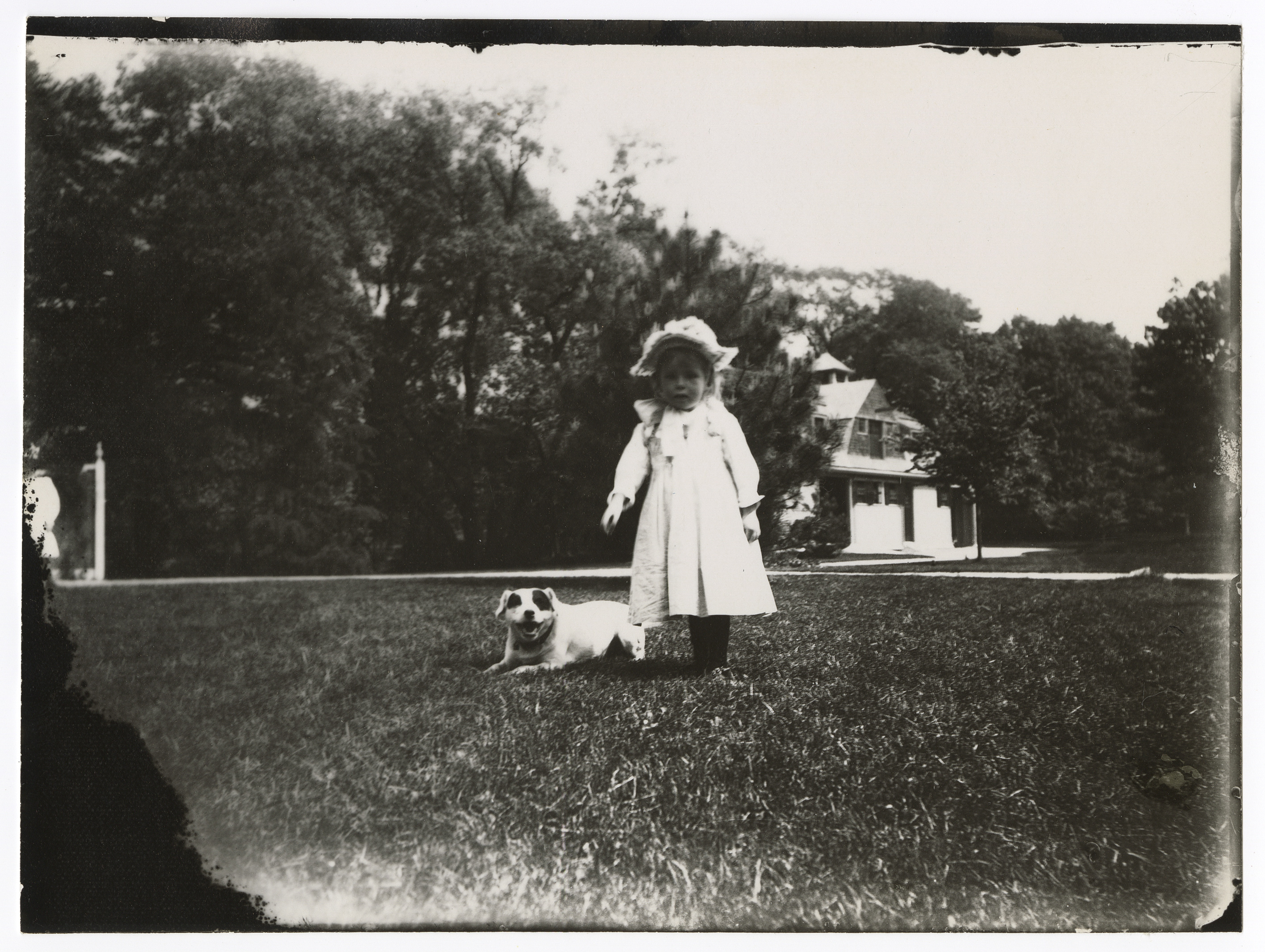 A white toddler in a hat and a long coat stands by a white dog with dark eye patches. Lawn in foreground, house and trees in background. Possibly an adult at left.