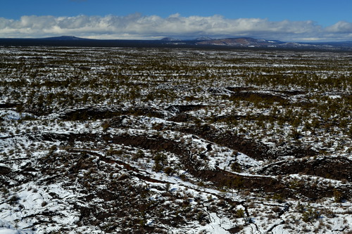 Viewed from above, snow dusts the surface of a winding, cracked surface of cooled lava flows.  Trees dot the surface, but the landscape's texture is still prominent.  Clouds line the sky above a distant mountain range.