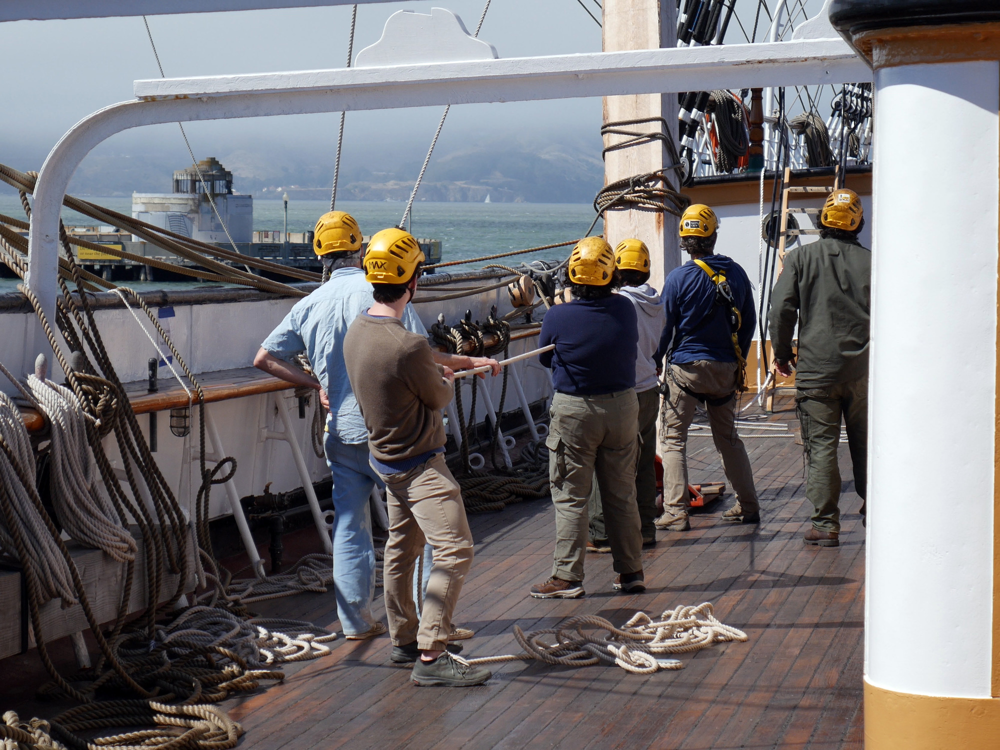 A group of volunteers and park staff wearing yellow hardhats works together to hold a line of rope on the deck of a ship