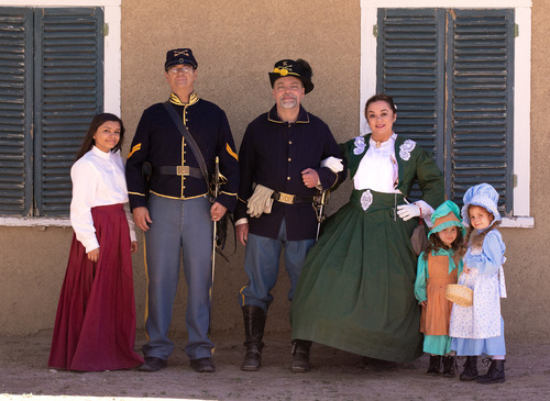 In historical 19th century dress, a soldier and his daughter are on the left, and another soldier with his wife and two little girls are on the right. They stand in front of an adobe fort wall with dark green shutters.