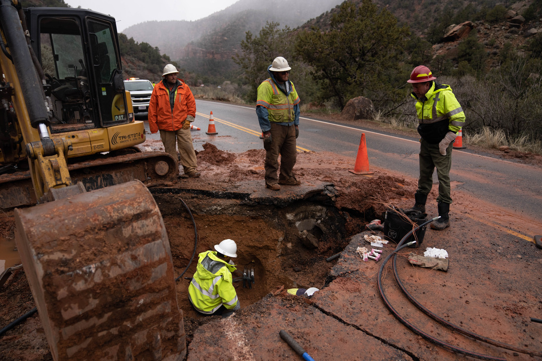 Four workers and electricians assess the damage a large rockfall caused on the road. One sits in a large hole caused by the rock.