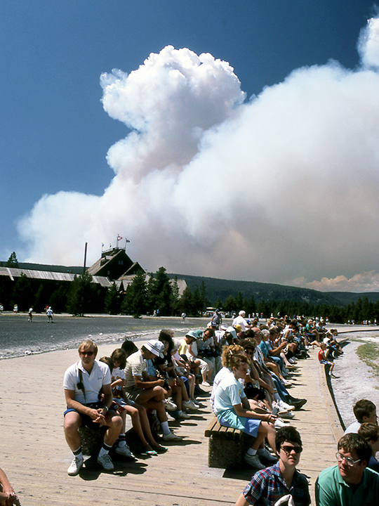 visitors waiting to see Old Faithful eruption with wildfire smoke in the distance on a sunny day