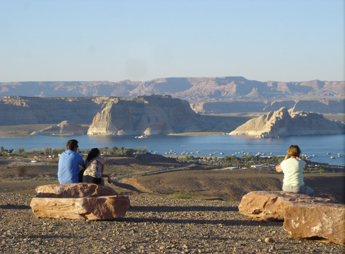 people sit on boulders facing away from the camera, looking at the view of Lake Powell