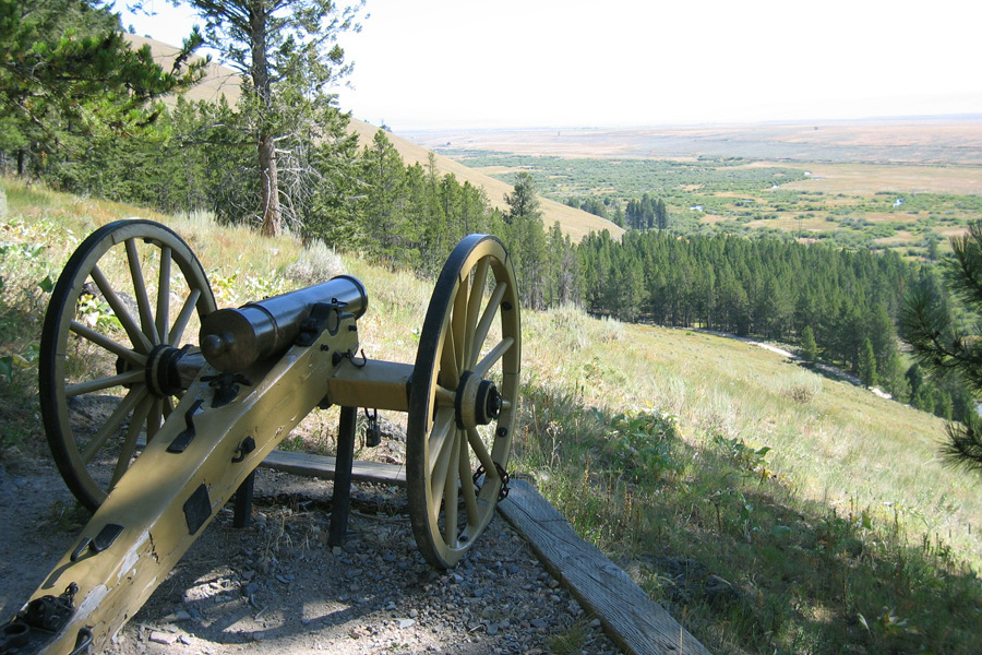 A view from behind a cannon includes a pine-covered hillside and a flat, open valley beyond.