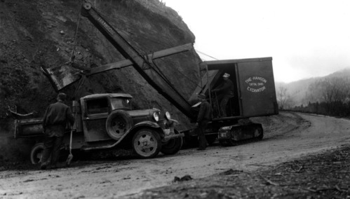 Historic photo of an excavator shovel cutting a slope along a road. A pickup truck is parked beside the shovel. 