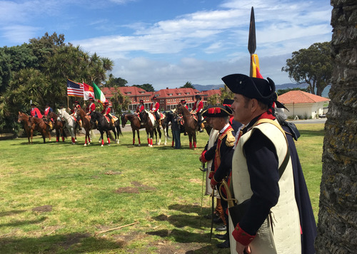 A line of men dressed in Spanish colonial military attire carrying Spanish, Mexican, and American flags on horseback stand on a grassy area in the San Francisco Presidio