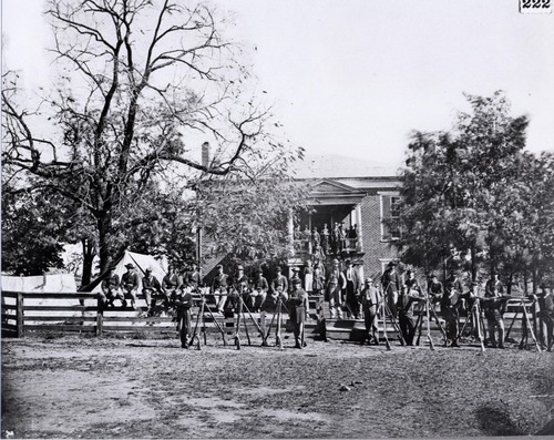 black and white photo of two story brick building with stairs to the second floor filled with soldiers posing. There are also soldiers standing and sitting by and on a fence surrounding the building