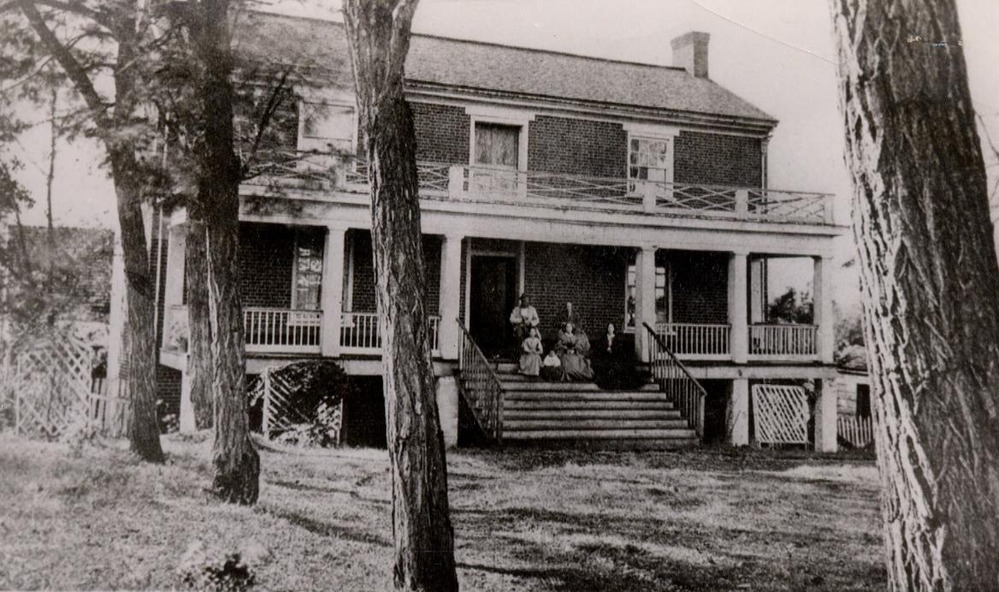 black and white photo of a 3 floor brick house with 4  trees in a row on the left of the house, the front of the house has a large porch and men and women are posing on the steps