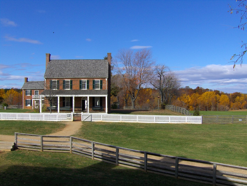 Clover Hill Tavern (1819) is a two story brick building with a porch across the front with a white fence across the front. Also included in the picture is the split rail fence which surrounds the courthouse. Behind the tavern are trees with autumn colored trees.