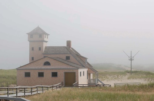 A lifesaving station building with a tall tower sits in the dunes shrouded in fog.