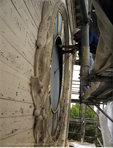 Color photo of a workman on scaffolding with his hands on a clock face much larger than himself.