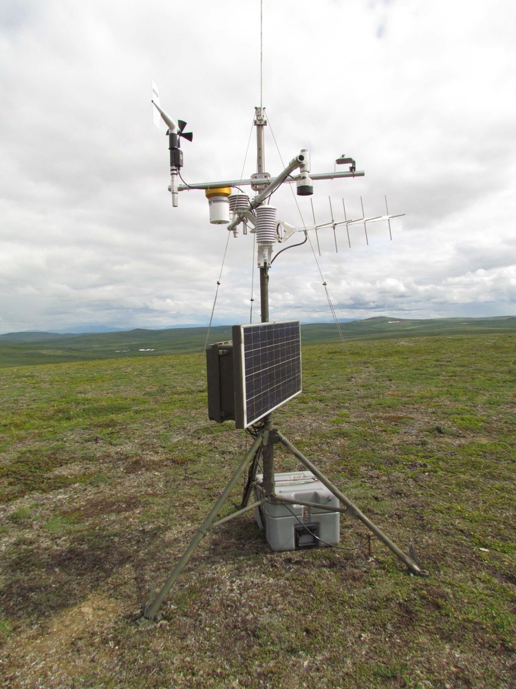 Solar panel, antenna, windmill, and collection containers on a tripod.