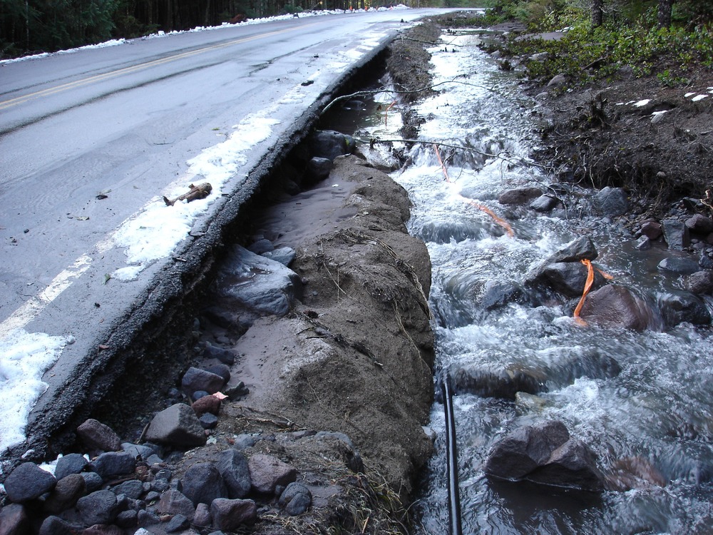 A creek flows along the edge of a road with the pavement undercut and piled with muddy debris. 