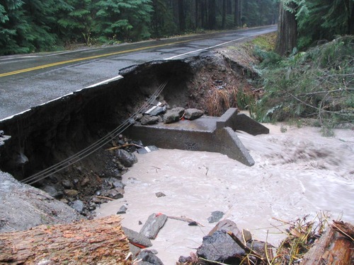 A paved road undercut by a creek, with the pavement hanging over an exposed concrete culvert. 
