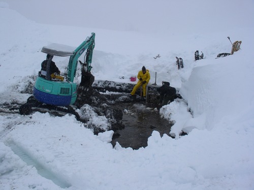 A backhoe and several people dig through snow to a muddy pool. 