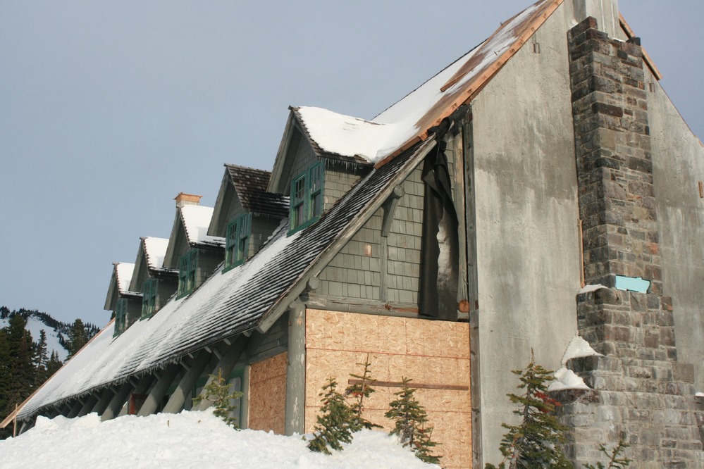 A large building with a steeply angled shingle roof. The side of the building is exposed, showing bare concrete and a stone chimney with the windows boarded up. 