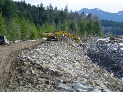 A large excavator on top of a levee moves large rocks along the side of the levee. 