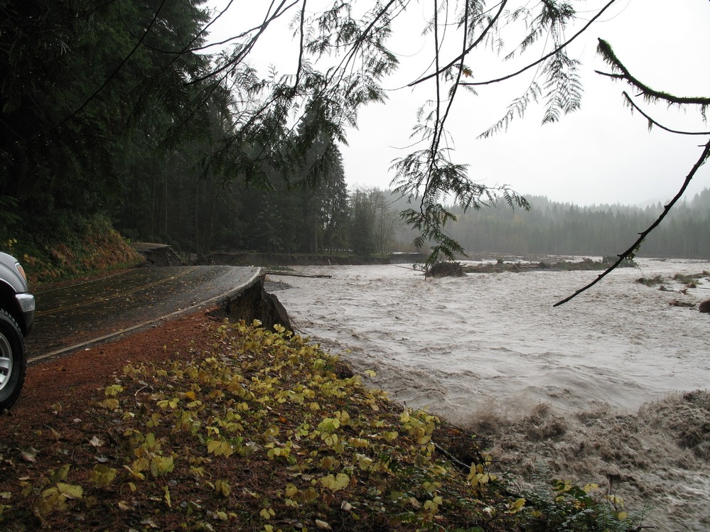 A wide muddy river flows next to a road that has been washed away. 