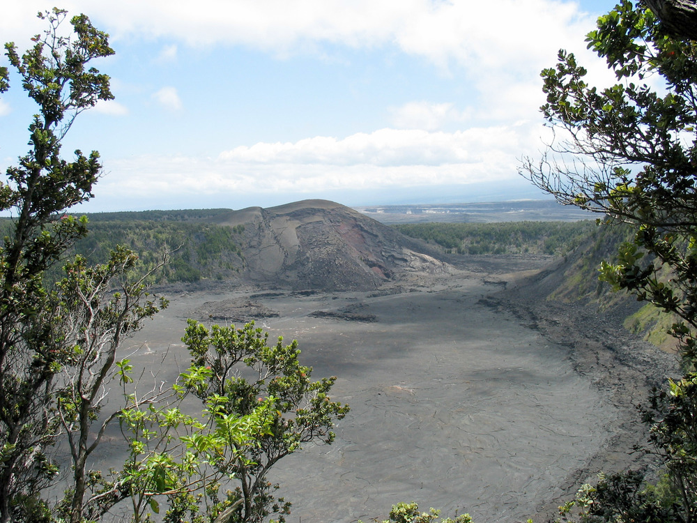 An expansive view from high above the flat, dark crater is framed by tree branches. 