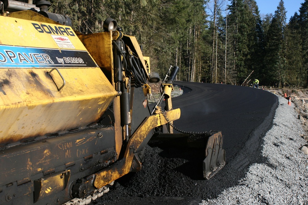 A yellow paving truck smoothes out asphalt along the edge of a road. 