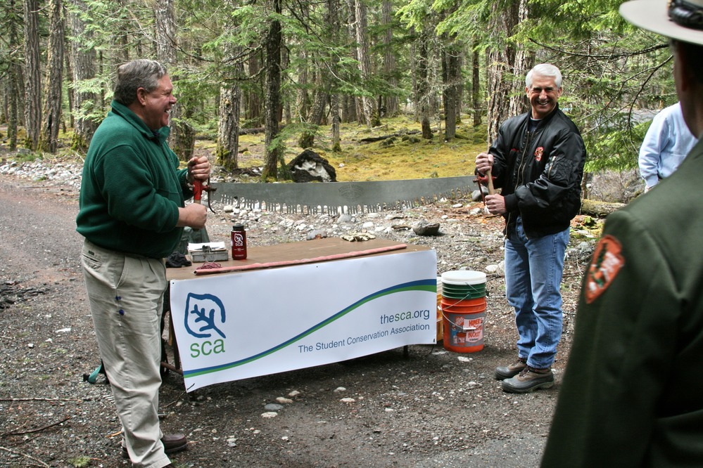 Two men hold up either end of a long tree saw in front of a table with a "SCA" sign hanging from it. 