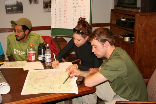 Three people sitting around a table look at a map and write in a notebook. 
