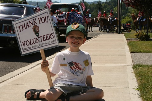 A smiling boy sitting on a sidewalk holds a "Thank you Volunteers" sign. 
