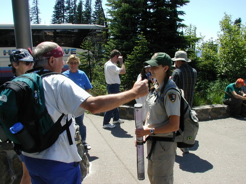 A volunteer talks to a group of people standing in a loose circle on a paved trail. 