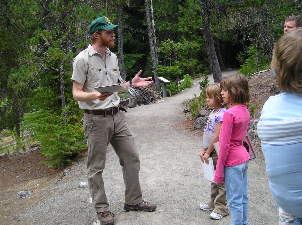 A man in a volunteer uniform stands along a gravel trail talking to several children. 