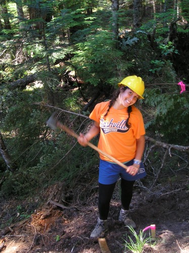 A smiling woman wearing a yellow hard hat swings a tool next to a trail. 