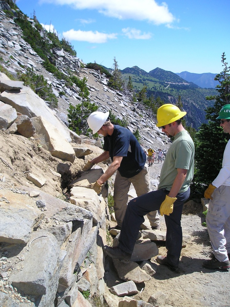 Several people wearing hard hats and gloves place a large rock into a rock wall along a trail. 
