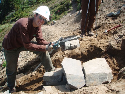 A volunteer wearing hard hats adjusts the placement of large rocks along a trail. 