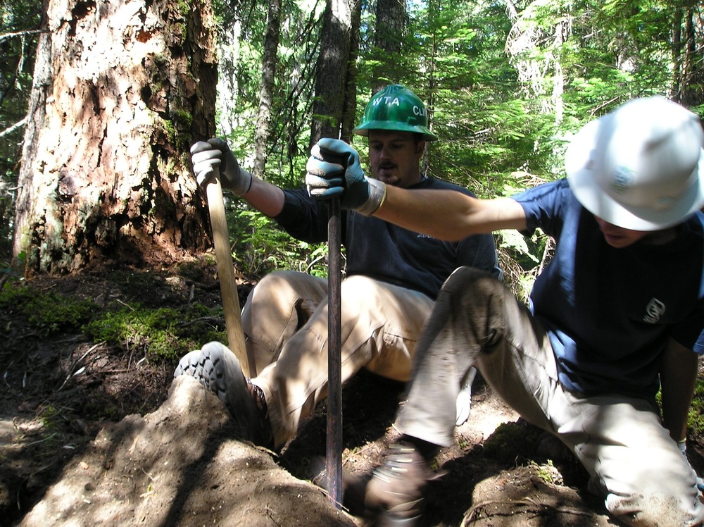 Two people use tools to pry up a rock along a section of trail. 