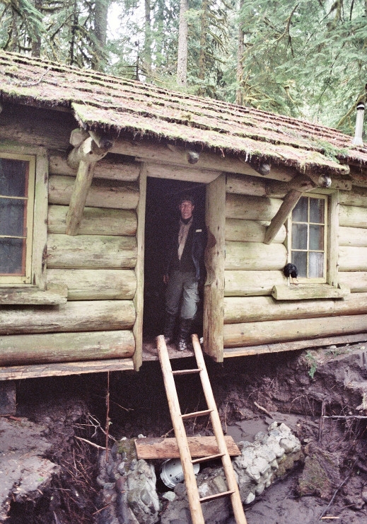 A ranger stands in the open doorway of a log cabin with a ladder going up to the doorway because a large river channel has washed out the ground under the cabin. 