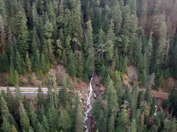 Aerial photo of a creek cutting through a forest and through a section of highway that is washed away.